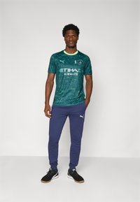 Groen voetbalshirt met patroon, korte mouwen en gele accenten, gecombineerd met marineblauwe joggingbroek en zwarte sneakers. Voorzien van logo's op het shirt en de broek.