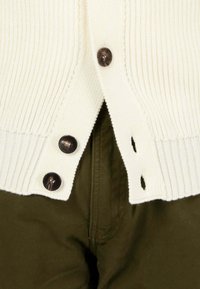 Cardigan a coste crema con bottoni marroni, caratterizzato da spacchi laterali e una finitura testurizzata, abbinato a pantaloni verde scuro.