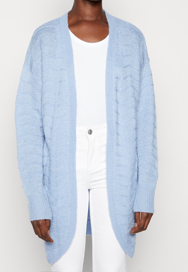 Hellblauer Strickcardigan mit offener Vorderseite, strukturiertem Muster, langen Ärmeln und gerippten Bündchen. Getragen über einem weißen T-Shirt und einer Hose.