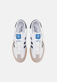 Een paar witte Adidas sneakers met marineblauwe strepen, een blauw logo op de tong, beige suède neuzen en rubberen zolen, gezien van bovenaf.