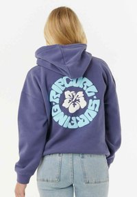 Lilla hættejakke med et stort hvidt hibiscus grafisk motiv og teal tekst på ryggen. Har en hætte med snor og en løs pasform, lavet af blødt stof.