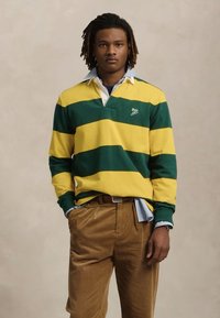 Jonge man met korte dreadlocks, gekleed in een geel en donkergroen gestreept rugbyshirt en een bruine corduroybroek, met zijn handen in zijn zakken, op een neutrale achtergrond.