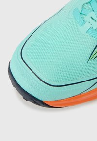 Světlé modré sportovní boty s texturovaným síťovaným svrškem, černými detaily a oranžovým panelem na patě. Mají zaoblenou špičku a zavazovací systém.