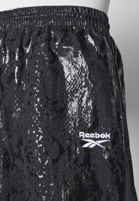Fekete sportos rövidnadrág fényes kígyómintás textúrával, rugalmas derékpánttal és fehér Reebok logóval a bal alsó részen.