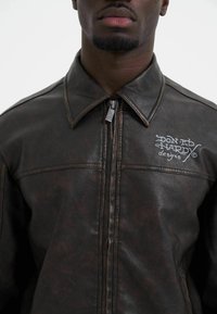 Brown leather jacket with a zip front, pointed collar, and visible stitching. Features embroidered logo on the left chest. Textured surface.