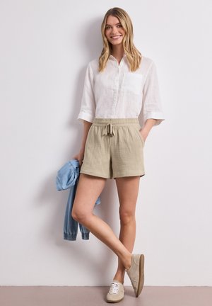 Femme souriante debout contre un mur, portant une chemise blanche à boutons, un short beige avec cordon de serrage, des baskets beiges, tenant une veste bleu clair dans la main gauche.