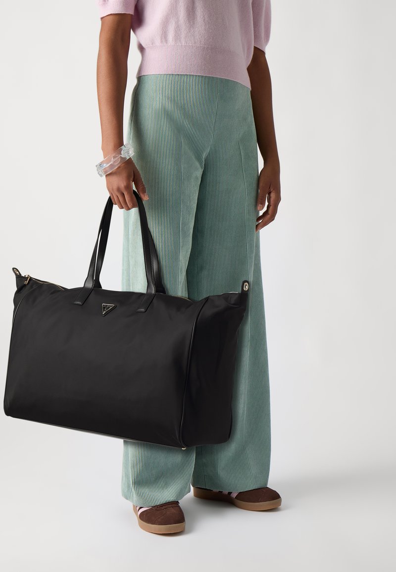 Sac fourre-tout en nylon noir avec des poignées en cuir, présentant un logo triangulaire et deux accents en métal argenté, montré avec un pantalon en velours côtelé vert clair.