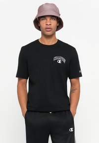 Mann iført en svart Champion t-skjorte, svarte shorts med logo, og en lilla bøttehatt, som står mot en enkel lys bakgrunn.