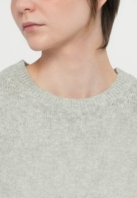 Fiatalember rövid barna hajjal, világosszürke kötött pulóvert viselve, balra nézve, semleges arckifejezéssel, fehér háttér előtt.