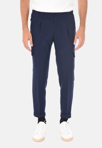 Pantaloni cargo blu navy con pieghe, vita elastica, tasche laterali e orli risvoltati. Abbinati a sneakers bianche su uno sfondo neutro.