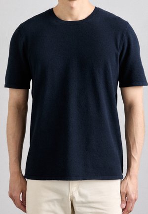 Mann trägt einen einfachen dunkelblauen kurzärmeligen Pullover und hellbeige Hose vor einem einfarbigen grauen Hintergrund.