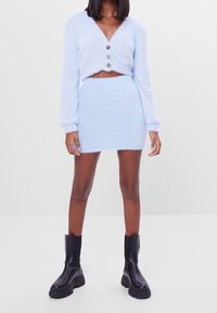 Cardigan corto peloso azzurro chiaro con tre bottoni, abbinato a una mini gonna coordinata. Il modello indossa stivali neri con tacco largo.