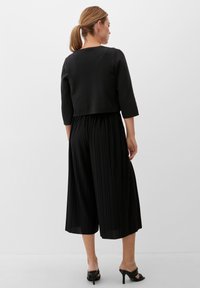 Vrouw met een paardenstaart, gekleed in een zwarte cropped sweater, geplooide wijde culottes en zwarte open teen hakken, staande tegen een witte achtergrond.