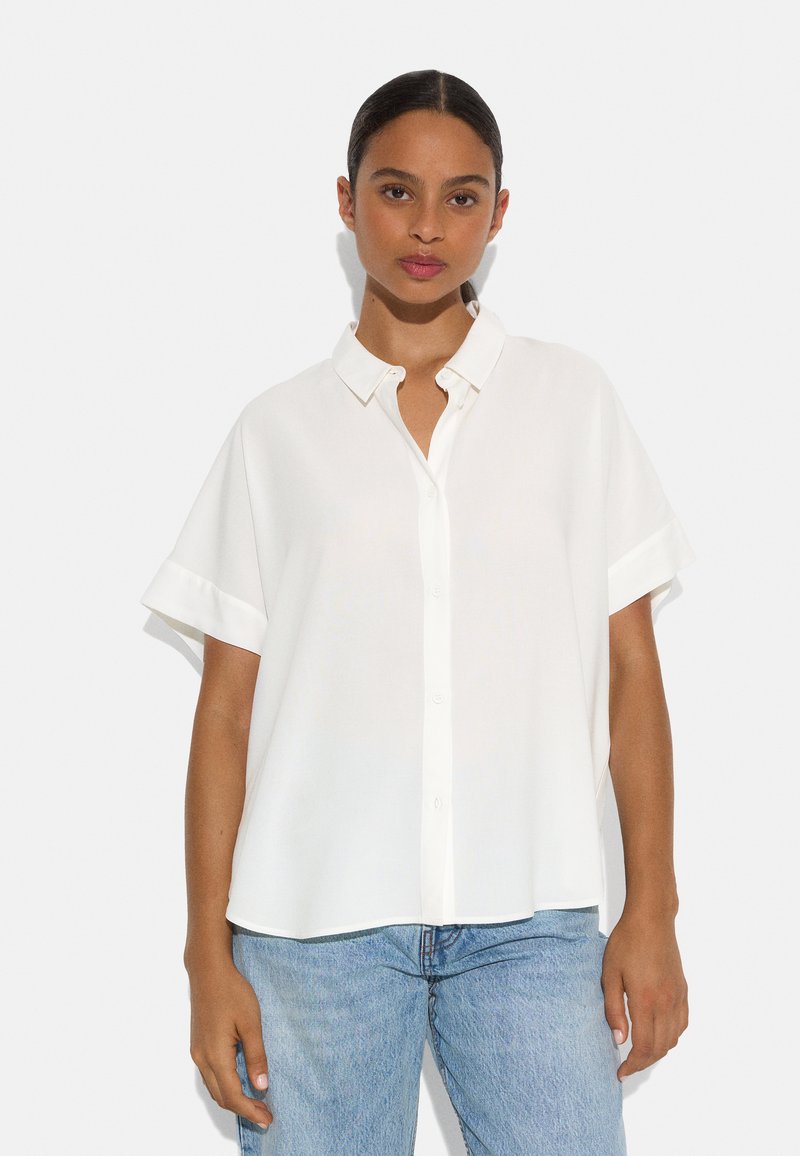 White short-sleeve blouse with a button-down collar, smooth fabric, and a relaxed fit, paired with light blue denim jeans.