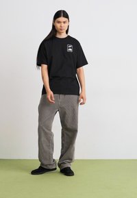 T-shirt en coton noir à manches courtes avec un logo sur la poche poitrine, associé à un pantalon large à rayures grises et des chaussures noires sur un sol vert.