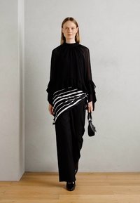 Kvinna som går, klädd i svart genomskinlig blus, svarta byxor, randig svart-vit scarf knuten i midjan, hållande en liten svart handväska.