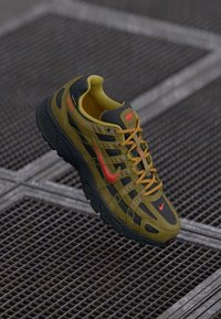 Nike apavi ar olīvzaļo un melno dizainu, ar teksturētu augšdaļu, izteiktiem sarkaniem akcentiem un izturīgu gumijas zoli.