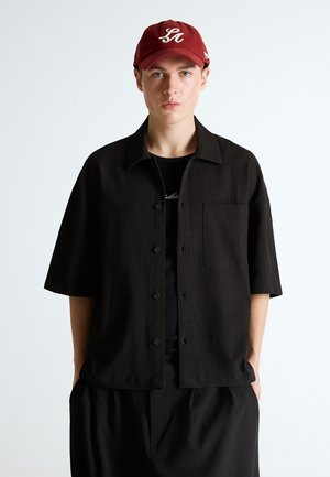 Mladić u crvenoj kapa s bijelim logotipom "LA" i širokoj crnoj košulji s kratkim rukavima stoji na jednostavnoj svijetloj pozadini.