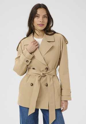 Trench coat bej cu curea, cu închidere dublă, epoleți la umeri și accente cu nasturi, purtat peste un top alb și blugi albaștri.
