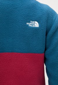 Chaqueta de forro polar azul y roja con tejido texturizado y logo bordado en blanco de "The North Face" en la parte superior de la espalda.
