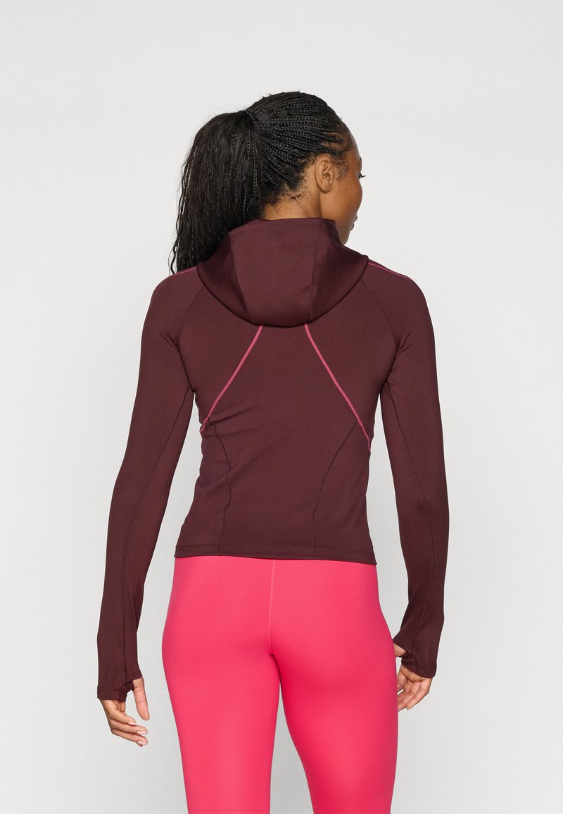 Maronenfarbener, taillierter Hoodie mit langen Ärmeln, Kapuze und pinken Akzentnähten. Kombiniert mit leuchtend pinken Leggings, die ein schlankes, sportliches Design zeigen.