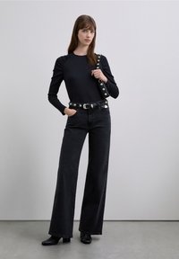 Black ribbed long-sleeve top with gathered shoulder detail, paired with high-waisted black wide-leg jeans and a studded black belt. Black heeled boots.