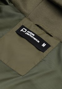 Interno della giacca verde oliva in tessuto liscio. Etichetta nera con la scritta "ombre performance" e taglia "M". Le cuciture sono pulite e ben rifinite.