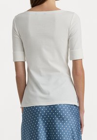 Vit, figurnära topp med trekvartslånga ärmar och vid halsringning. Slät textur, med uppvikt manschetter och en låg bakre fåll.