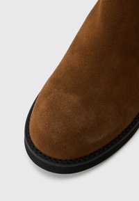 Καφέ σουέτ ankle boot με στρογγυλή μύτη, μαύρη καουτσούκ σόλα και ορατές ραφές κατά μήκος της άκρης. Η υφή είναι απαλή και λεία.