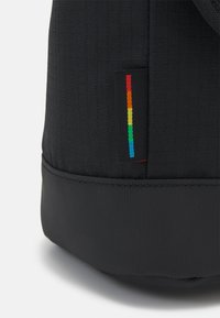 Sac noir avec une surface texturée, doté d'une étiquette colorée en arc-en-ciel et d'une section inférieure noire lisse, mettant en avant un design minimaliste.