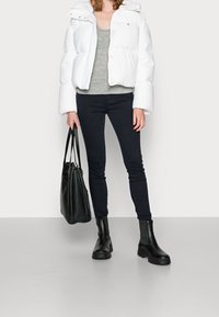 Biała puchowa kurtka z kapturem, szara bluzka, czarne rurki i czarne botki za kostkę. Nosi czarną torbę typu tote. Gładka faktura, casualowy styl.