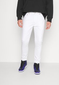 Bílé džíny slim fit, s pěti kapsami, poutky na opasek a jemnou strukturou. Spárovány s černými a fialovými vysokými teniskami.