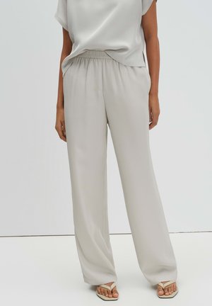 Vrouw draagt losse, lichtbeige broek met elastische tailleband en bijpassende top, gecombineerd met beige teenslippers, staand tegen een witte achtergrond.