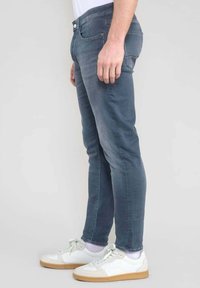Jeans de corte slim para hombre en denim gris oscuro desgastado, con un diseño estándar de cinco bolsillos, cremallera y cierre de botón. Combinados con zapatillas blancas.