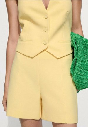 Gul ærmeløs knappet vest med matchende højtaljede shorts, person holder en tekstureret lysegrøn clutch under armen.