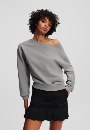 Pelēks plecu atklājošs džemperis ar garām piedurknēm, ribotām aprocēm un apakšmalu, uz kura melnā krāsā ir uzraksts "KARL LAGERFELD JEANS". Kombinēts ar melnu plisēto svārku.