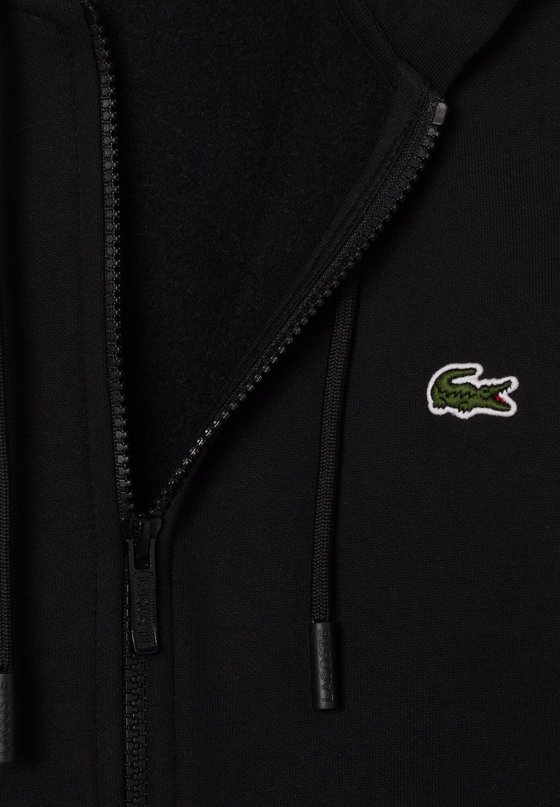 Haut à fermeture éclair noir avec intérieur texturé, cordons et logo de crocodile vert brodé sur la poitrine gauche. Fermetures éclair et quincaillerie noires.