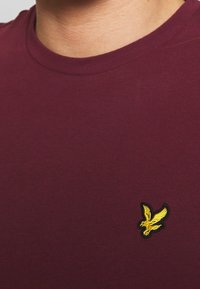 Bordová tričko vyrobené z bavlny, s kulatým výstřihem, na levé straně má malé žluté vyšívané logo orla.