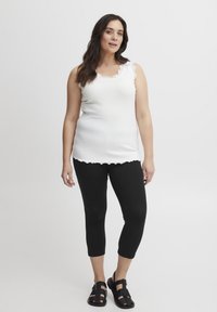 Top in cotone bianco a coste con bordi ondulati, abbinato a leggings neri a tre quarti e sandali neri con cinturini. Materiali lisci e design aderente.