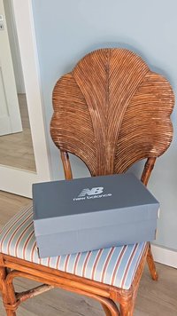 Scatola grigia di New Balance su una sedia intrecciata con uno schienale a forma di foglia. Il cuscino a righe blu e bianco aggiunge contrasto.