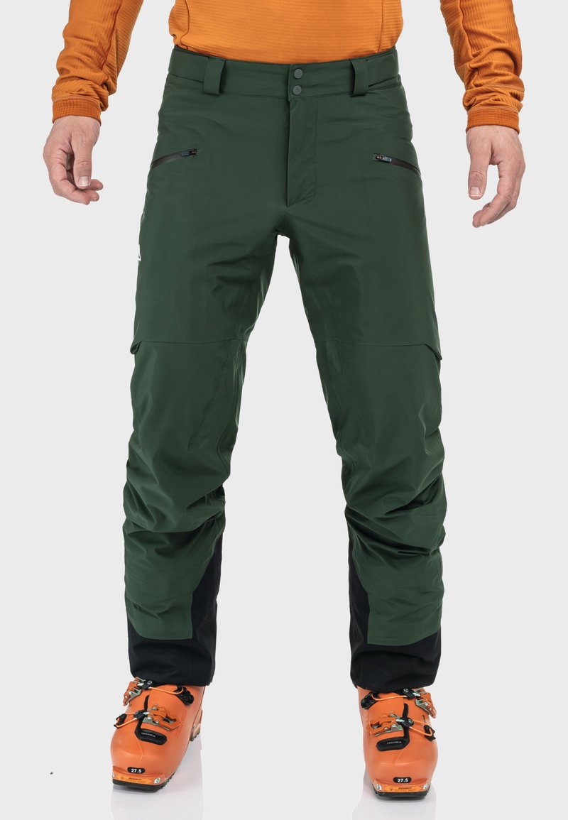 Grüne Outdoor-Hosen mit wasserabweisender Beschichtung, ausgestattet mit Reißverschlusstaschen und schwarzen Akzenten, getragen mit orangefarbenen Skischuhen.