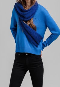 Niebieski sweter dzianinowy z okrągłym dekoltem, zestawiony z teksturowanym niebieskim szalikiem. Czarne jeansy o dopasowanym kroju.