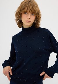 Marineblauer Rollkragenpullover mit strukturierter Oberfläche und erhabenen Perlenakzenten, mit langen Ärmeln und gerippten Manschetten, vor einem neutralen Hintergrund.