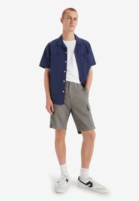 Camicia azzurra a maniche corte con bottoni e due tasche frontali, abbinata a pantaloni cargo grigi. Una t-shirt bianca e delle sneakers completano l'outfit.