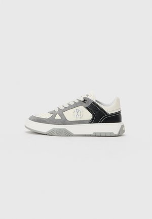 Zapatilla baja en blanco, ante gris y cuero negro con cordones blancos y bordado del logo en el lateral, exhibida sobre un fondo simple.