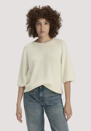 Frau mit lockigem Haar, die einen lockeren cremefarbenen Strickpullover mit ellenbogenlangen Ärmeln und blaue Jeans trägt, vor einem schlichten Hintergrund stehend.