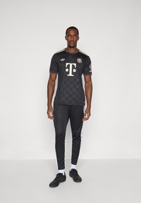 Jersey deportivo negro con patrón de tablero de ajedrez, acentos dorados y logo. Combinado con pantalones negros con rayas laterales blancas y zapatillas deportivas.