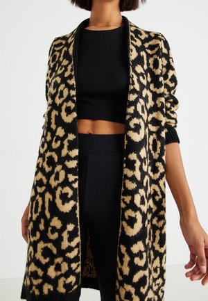 Mujer con un cárdigan largo abierto con estampado de leopardo sobre un top corto negro y pantalones negros de talle alto, de pie sobre un fondo blanco.