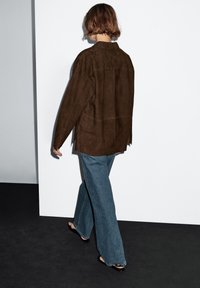 A person with short hair walks away, wearing a brown suede shirt and flared blue jeans, against a minimalist white wall.