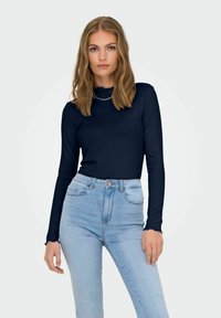 Top lungo a maniche corte a coste color navy con collo alto e polsini arricciati, abbinato a jeans chiari a vita alta. Design semplice, texture morbida.